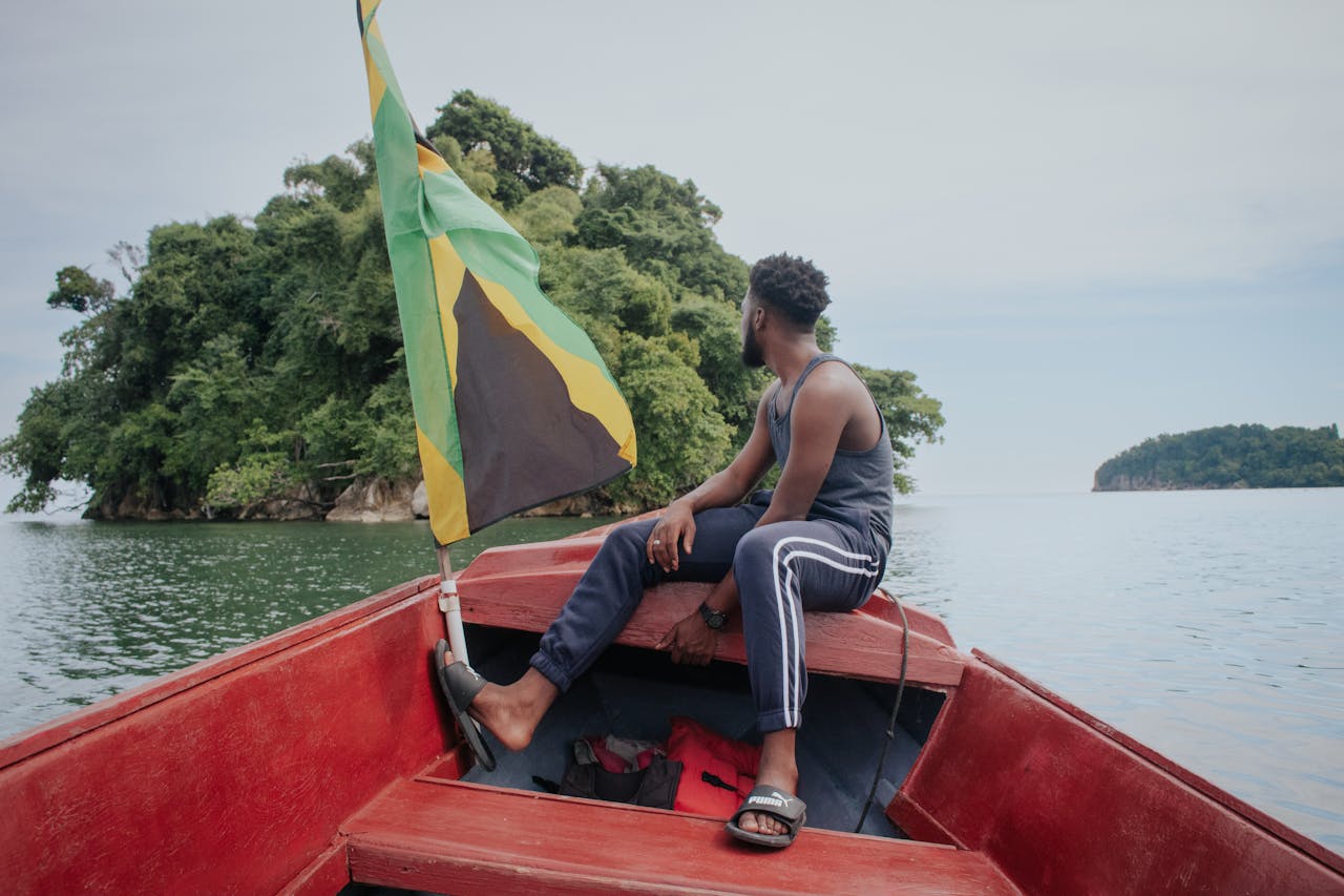 Man enjoying a peaceful boat ride near a lush island with a Jamaican flag.